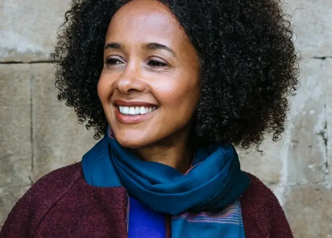 A portrait of Black novelist and journalist Diana Evans. She has shoulder length hair and is wearing a maroon jacket, blue shirt and blue scarf.