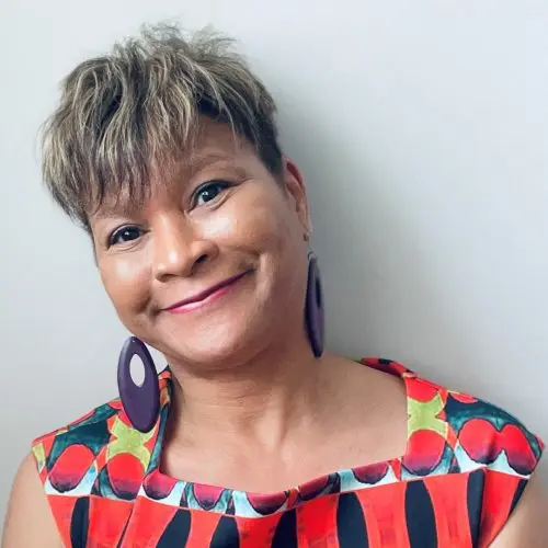 A portrait of Black writer for theatre, tv and radio and children's author Trish Cooke. She has short mid-brown hair, is wearing large purple earrings and a short-sleeved, square-necked patterned top in red, blue and yellow.