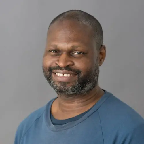 A portrait of the Black writer and academic Oladipo Agboluaje. He has close cropped hair and a greying beard and is wearing a blue sweatshirt.