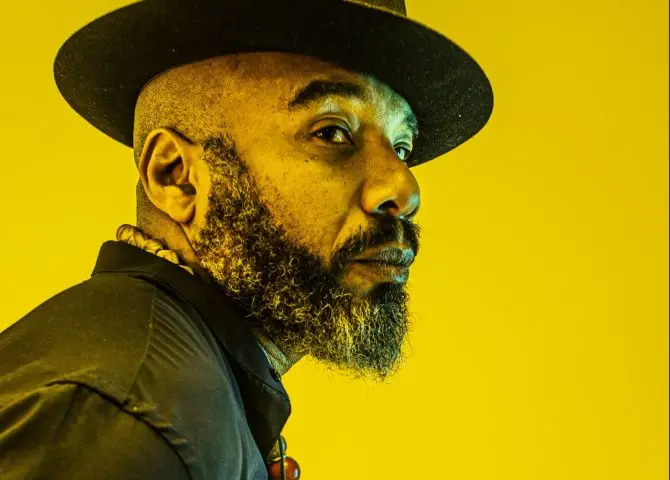 An image of Caribbean writer Anthony Joseph. He is wearing a black trilby, black shirt and is bearded. This image is a side profile and features a yellow background.