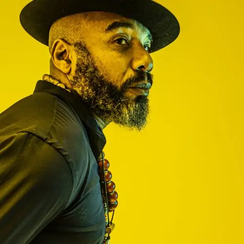 Writer Anthony Joseph. A bearded man pictured from the waist up wearing a black hat, black shirt, beaded necklace pictured against a yellow background.