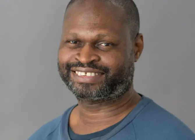 A portrait of the Black writer and academic Oladipo Agboluaje. He has close cropped hair and a greying beard and is wearing a blue sweatshirt.
