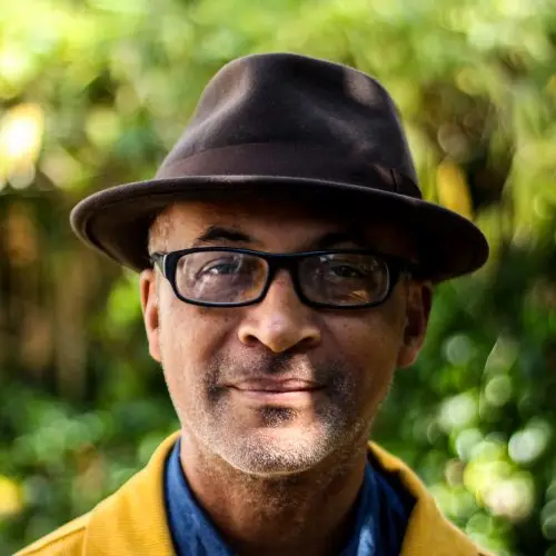 An image of the Black writer Colin Grant taken outside. He is wearing glasses and is sporting a brown hat, blue shirt and yellow jacket.