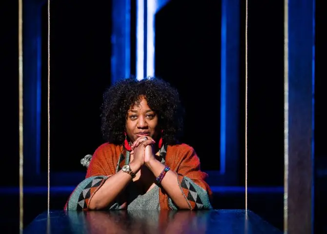 The Black female author Juliet Gilkes Romero is pictured sitting with her arms on a table, chin resting on her clasped hands. She is wearing an orange shawl with blue, black and grey detail and a grey v-neck top