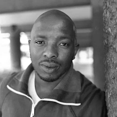A portrait of Black poet Nick Makoha. He is pictured outside leaning against a wall. He's wearing a white t-shirt and tracksuit top.