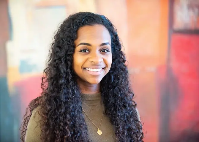 A portrait of the writer Hafsa Zayyan. She has long curly hair and is wearing a khaki long sleeved top and gold necklace.