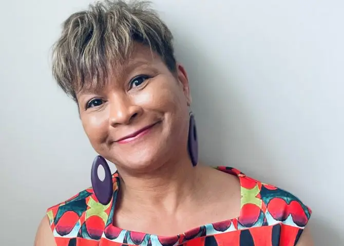 A portrait of Black writer for theatre, tv and radio and children's author Trish Cooke. She has short mid-brown hair, is wearing large purple earrings and a short-sleeved, square-necked patterned top in red, blue and yellow.