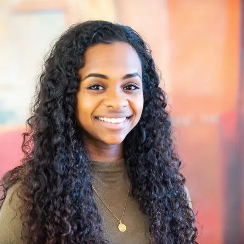 A portrait of the writer Hafsa Zayyan. She has long curly hair and is wearing a khaki long sleeved top and gold necklace.